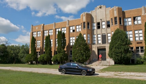A black car parked in front of a large, abandoned brick building with trees and a person standing on the steps.