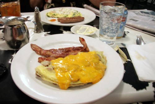 A plate with a cheese omelet, bacon, and a glass of water, with another plate of food in the background.