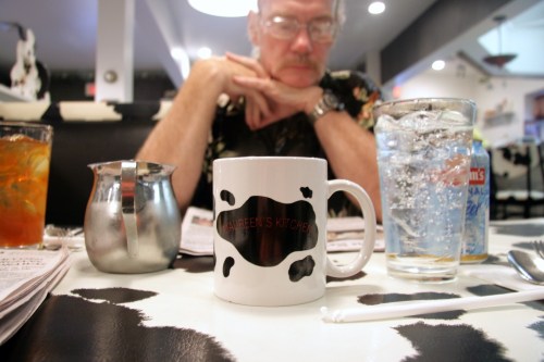 A coffee mug with "Maureen's Kitchen" sits on a table, with a man in the background and drinks nearby.