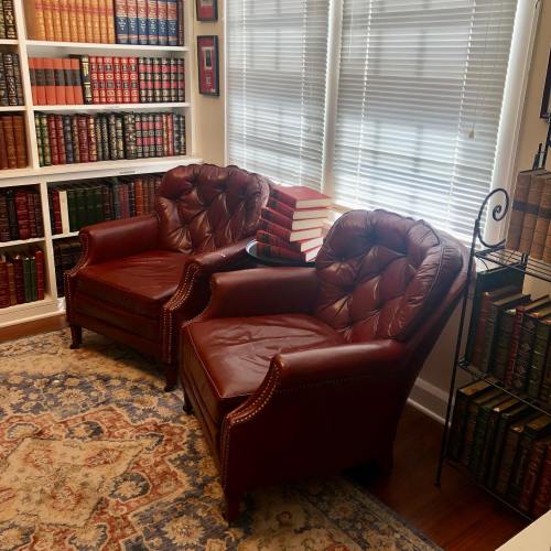 Two red leather armchairs in a cozy library with bookshelves filled with books and a patterned rug.