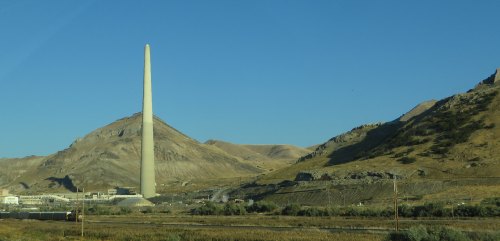 A tall, slender smokestack stands against a clear blue sky, surrounded by dry hills and industrial buildings.