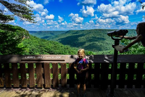 A girl stands at a scenic overlook with mountains and a blue sky in the background, enjoying the view.