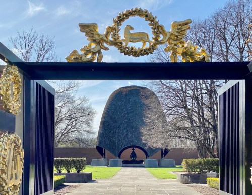 A decorative gate frames a modern, curved building surrounded by trees and manicured landscaping under a clear blue sky.