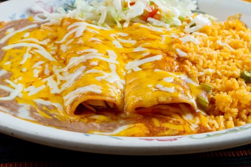 A plate of cheesy enchiladas topped with sauce, served with rice and shredded lettuce on the side.