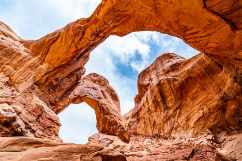 Two natural rock arches framed by towering red rock formations under a blue sky with scattered clouds.