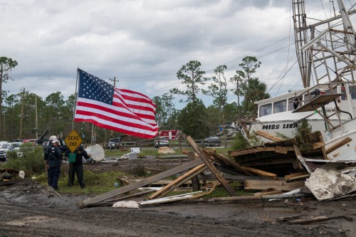 A tattered American flag waves near a damaged boat and debris in a storm-affected area, with responders in the background.