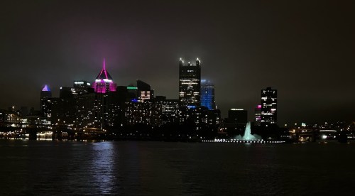 City skyline at night with illuminated buildings and a fountain, reflecting on the water.