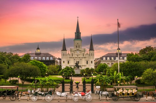 A scenic view of Jackson Square in New Orleans at sunset, featuring horse-drawn carriages and St. Louis Cathedral.