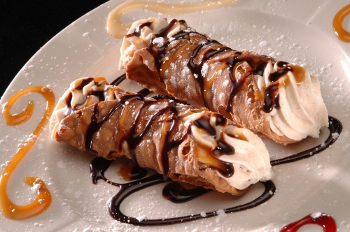 Two chocolate-filled cannoli drizzled with chocolate and caramel sauce, garnished with whipped cream and powdered sugar.