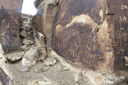 Petroglyphs etched into rock walls, surrounded by boulders and a dirt path in a rugged outdoor setting.
