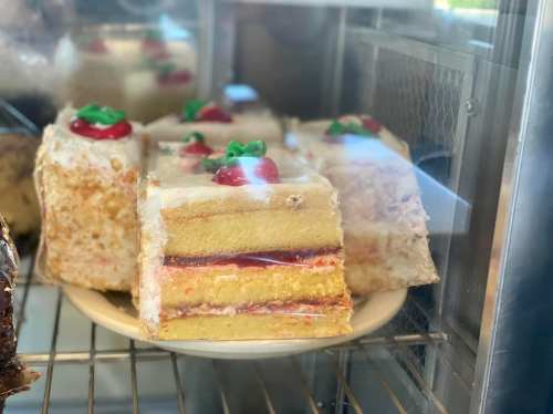 A slice of layered cake with frosting, topped with a cherry and green decoration, displayed in a glass case.