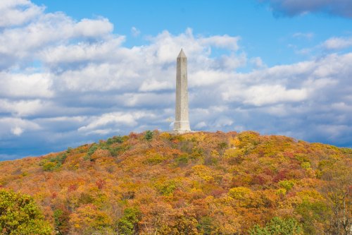 A tall monument stands atop a hill surrounded by vibrant autumn foliage under a partly cloudy sky.