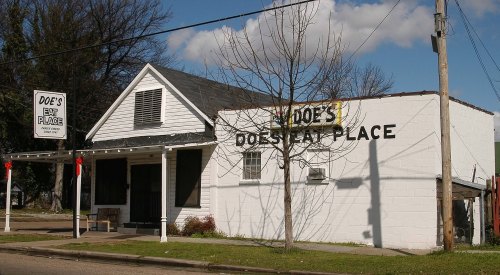 A white building with a black sign reading "Doe's Eat Place," featuring a porch and trees in the background.