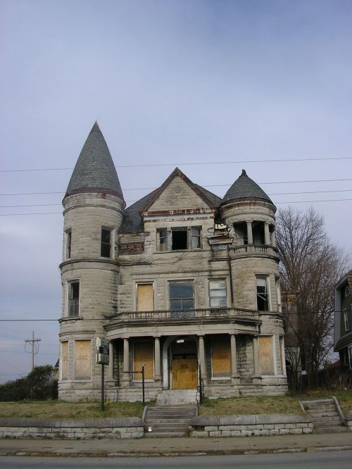 A dilapidated Victorian-style mansion with a turret, boarded windows, and overgrown grass under a cloudy sky.