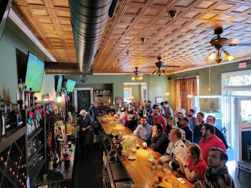A lively bar scene with a crowd watching sports on TV, seated at the bar and tables, with festive decorations.