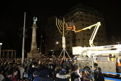 Crowd gathered at night for a menorah lighting ceremony, with a cherry picker lifting the menorah into place.