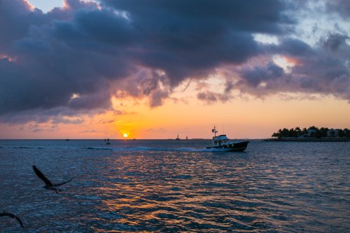A boat navigates the water at sunset, with colorful clouds and silhouettes of distant boats and land in the background.
