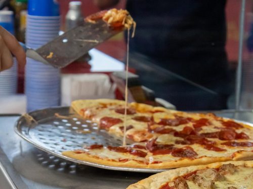 A hand lifts a slice of pepperoni pizza, with cheese stretching from the pie on a metal tray.