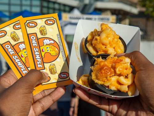 Two hands hold a container of cheesy macaroni and cheese with two sample tickets in the background.