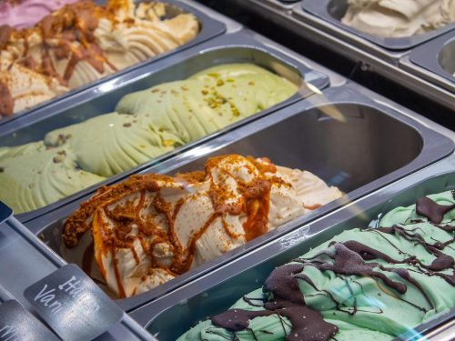 A variety of gelato flavors in metal containers, including pistachio, vanilla, and chocolate drizzled with sauce.