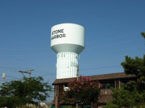 A light blue water tower labeled "STONE HARBOR" stands against a clear blue sky, surrounded by trees and a building.
