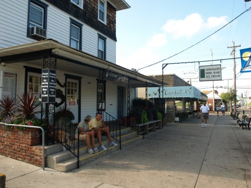 A street view of a small town with shops, featuring people sitting on steps and a sign for an ice cream shop.