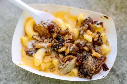 A serving of macaroni and cheese topped with crispy bits of meat and fried onions in a white container.