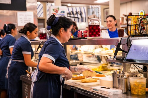 A busy kitchen scene with women in blue uniforms preparing food and serving customers at a counter.