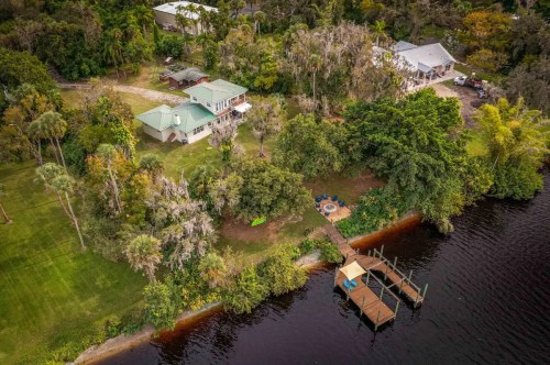 Aerial view of a riverside property with a house, dock, and seating area surrounded by lush greenery.