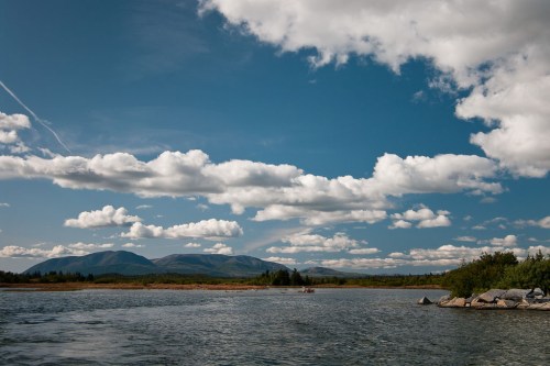 A serene river scene with mountains in the background and fluffy clouds in a blue sky.