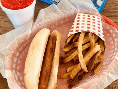 A hot dog in a bun next to a container of crispy French fries and a cup of red beverage on a wooden table.