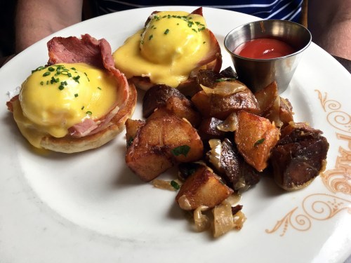 A plate of eggs Benedict with ham and hollandaise sauce, served with crispy potatoes and a small cup of ketchup.