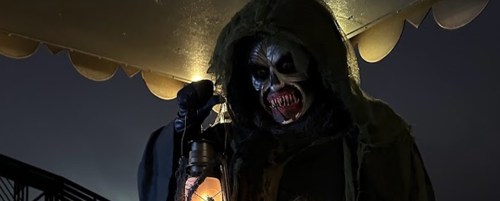 A sinister figure in a hooded cloak holds a lantern, revealing a terrifying skull-like face in a dark setting.
