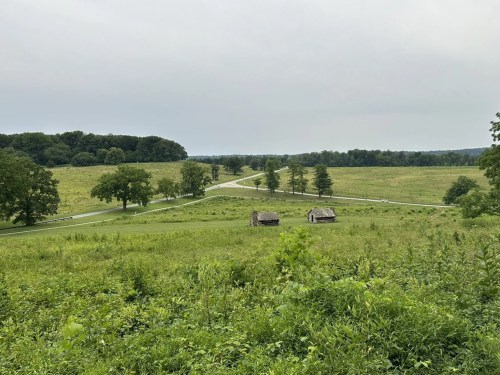 A scenic view of a green landscape with two small cabins, winding road, and trees under a cloudy sky.