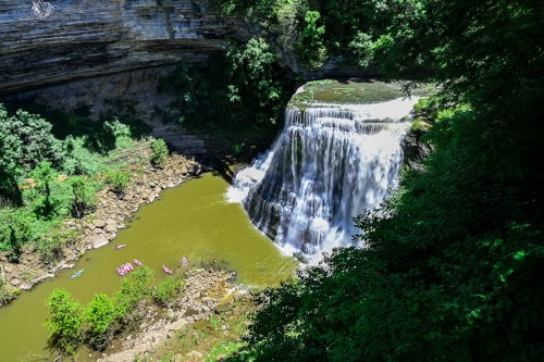 A scenic waterfall cascades into a green pool, surrounded by lush trees and rocky cliffs, with kayakers below.