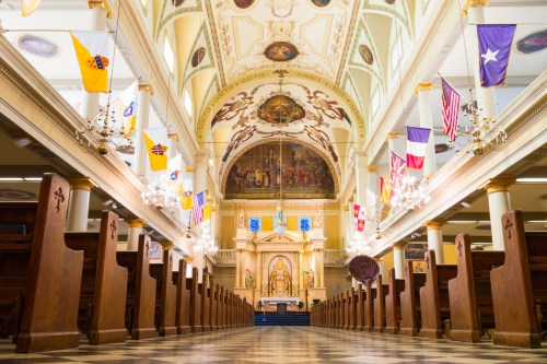 Interior of a grand church featuring ornate ceilings, wooden pews, and colorful flags hanging from the walls.