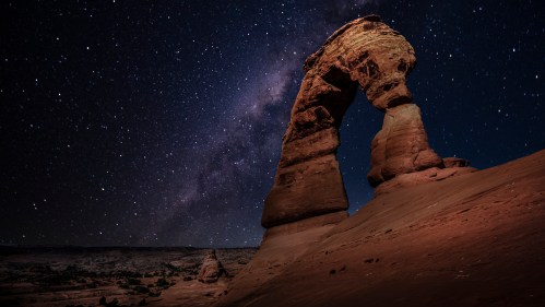 A stunning night sky filled with stars above a large rock formation, resembling an arch, in a desert landscape.