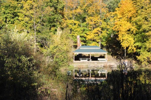 A wooden pavilion surrounded by colorful autumn trees, reflecting in a calm pond.