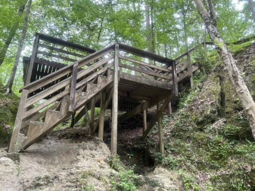 Wooden staircase with multiple levels, surrounded by lush green trees and rocky terrain.