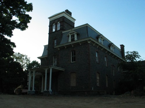 A large, historic stone house with a tower, featuring a porch and surrounded by trees at dusk.
