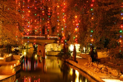 A serene river scene at night, adorned with colorful holiday lights on trees and a bridge reflecting in the water.