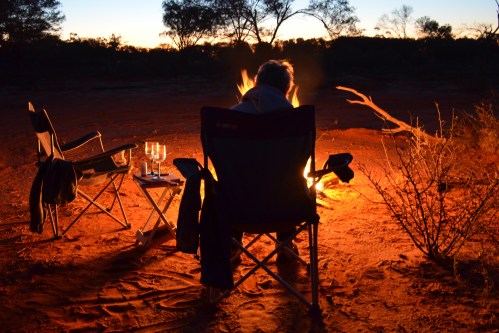 A person sits by a campfire in the desert at sunset, with chairs and glasses nearby, surrounded by nature.