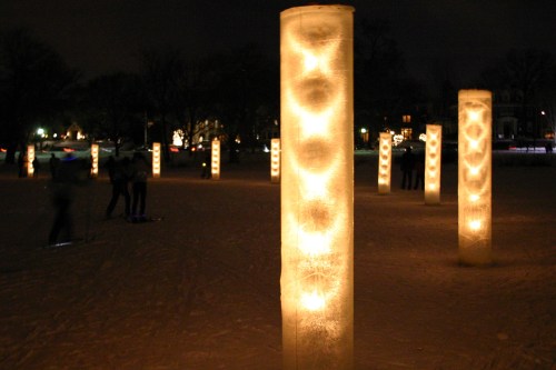 Illuminated ice columns in a snowy park at night, with blurred figures in the background enjoying the winter scene.