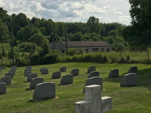 A cemetery with gray tombstones in the foreground and a dilapidated building surrounded by greenery in the background.