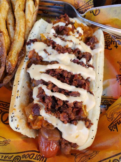 A loaded hot dog topped with chopped meat, onions, and drizzled with sauce, served with crispy fries on the side.