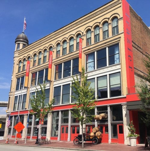 Historic building with large windows, red accents, and a vintage car parked outside, surrounded by trees.