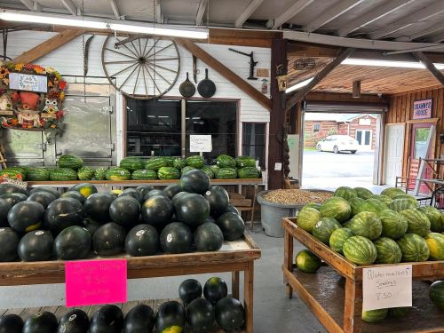 A market interior with tables of watermelons and signs for prices, featuring a rustic decor and open doorway.