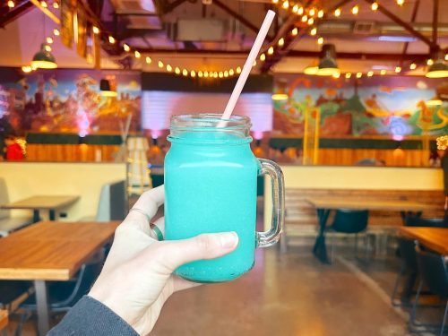 A hand holding a blue drink in a mason jar with a straw, set against a vibrant, illuminated restaurant interior.