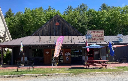 A rustic snack shack with a wooden exterior, colorful flags, and outdoor seating, surrounded by green trees.
