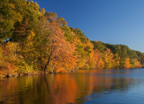 A serene lake surrounded by vibrant autumn foliage reflecting on the water under a clear blue sky.